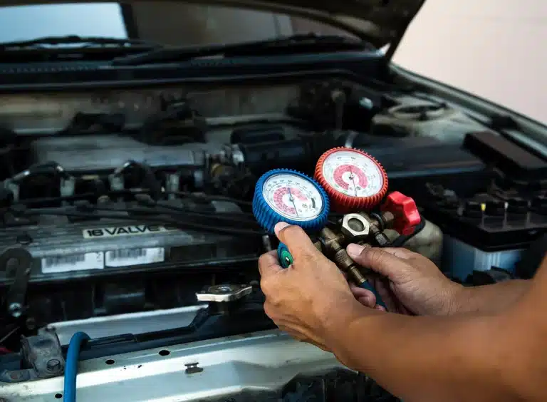A person uses a set of pressure gauges to check or service the air conditioning system under the hood of a car with the engine exposed.