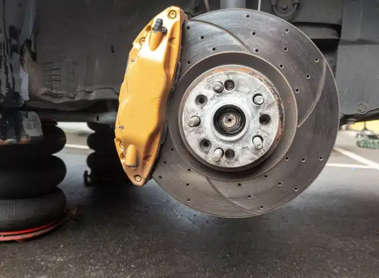 A close-up view of a car’s brake disc and gold brake caliper, mounted on the wheel hub, with the wheel removed. The car is elevated, exposing the suspension and braking components.