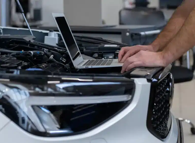 A person uses a laptop placed on the raised hood of a car, likely diagnosing or servicing the vehicle. The cars engine is partially visible, and the setting appears to be a workshop or garage.
