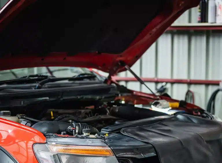 A red car with its hood open in a garage, exposing the engine bay for maintenance or repair. The car is covered with a black protective mat on the front fender. Shelves and tools are visible in the background.