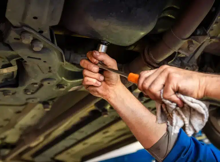 A mechanic’s hands use a wrench and screwdriver to work under a car, possibly changing the oil or repairing a part. One hand holds a cloth, and the underside of the vehicle is visible above.
