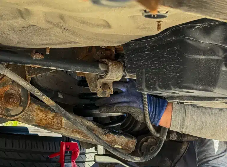 A person wearing a blue glove works under a vehicle, handling parts of the suspension system near a black fuel tank. A tire, rusted metal, and a red jack stand are also visible.