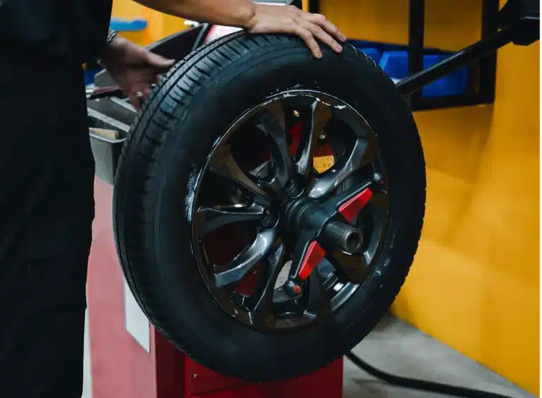 A person is placing a car wheel with a black tire and alloy rim onto a machine in an auto repair shop with yellow walls in the background.