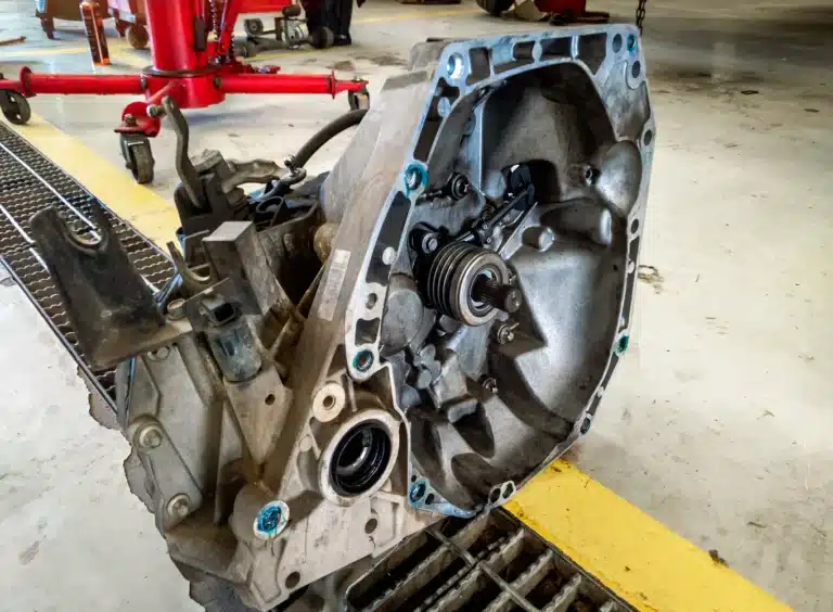 A close-up of a car transmission removed from a vehicle, resting on a garage floor near a drainage grate. The transmission case is open, revealing internal components and gears. Red automotive equipment is visible in the background.