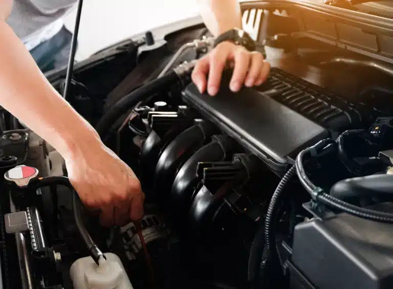 A person’s hands are working on a car engine, adjusting components under the hood with tools. The engine parts and wires are visible in close-up.