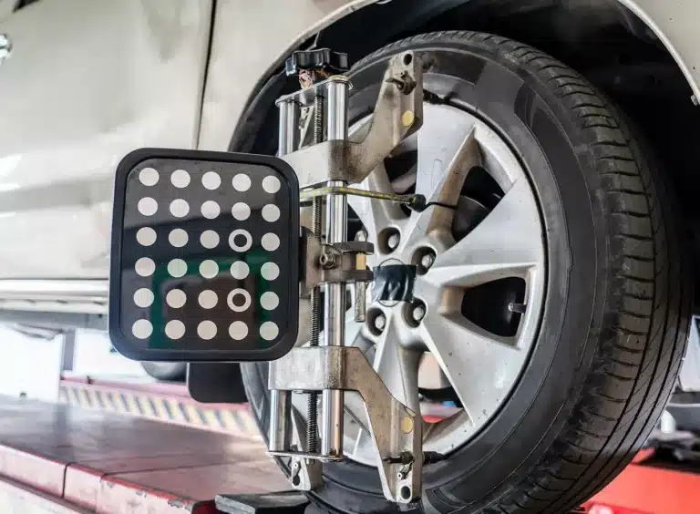 A car wheel with an alignment tool attached, featuring a metal frame and a black panel with white dots, during a wheel alignment procedure in an auto repair shop.