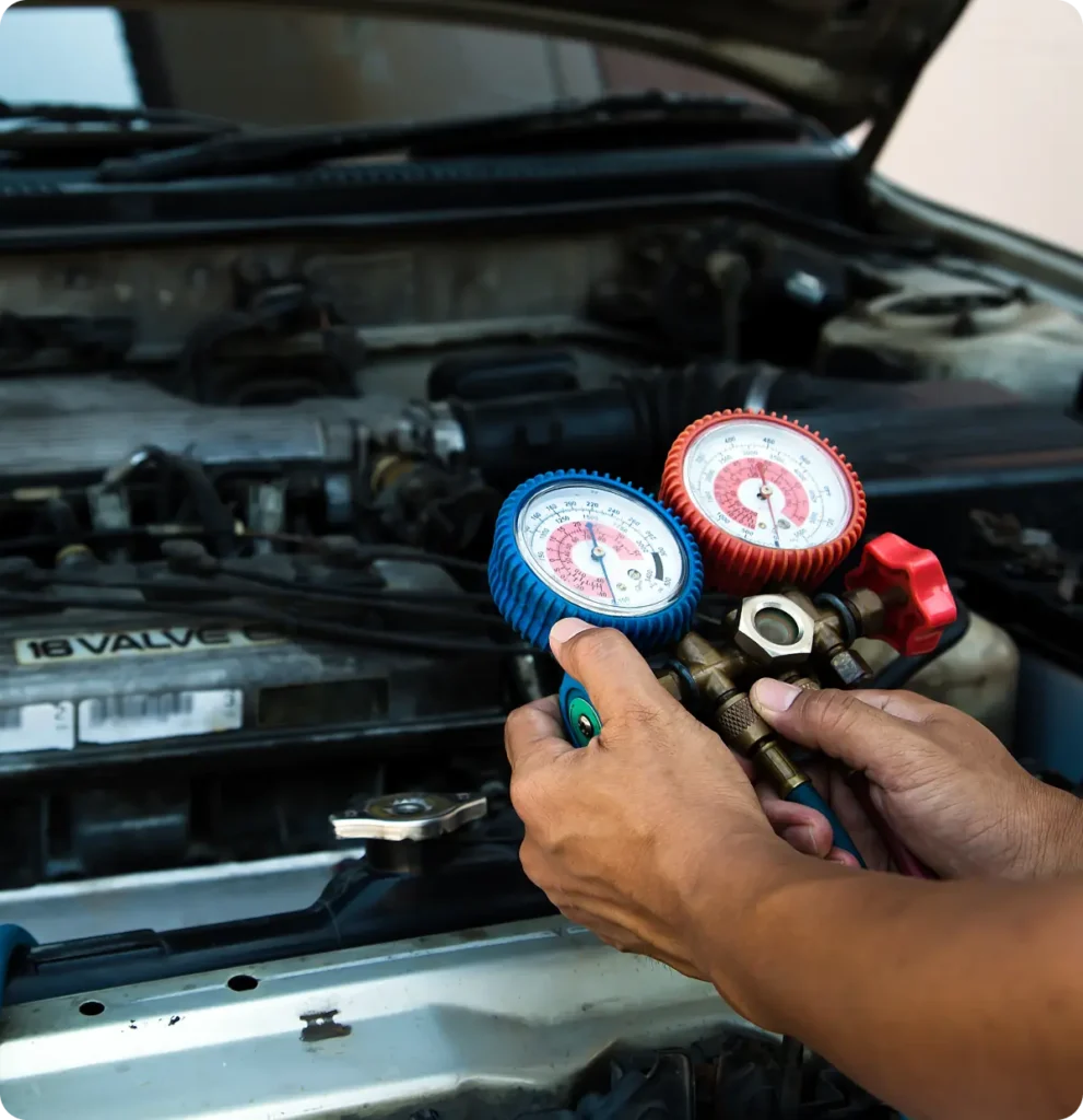 A person checks a car engine using a manifold gauge set with red and blue dials, likely to measure or service the air conditioning system. The car hood is open, revealing the engine.