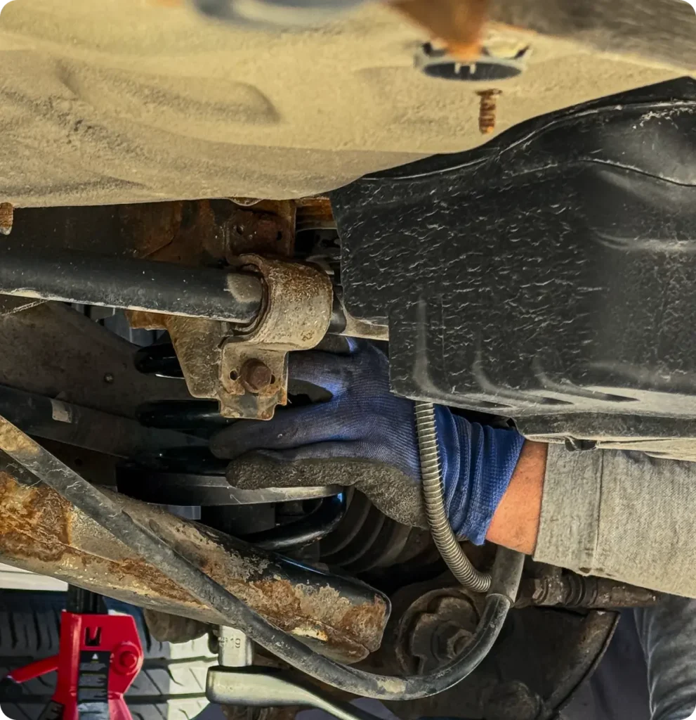 Steering & suspension repair, auto repair in Springfield, OR, at Absolute Air & Auto Repair. A person wearing a blue work glove repairs or inspects the underside of a vehicle, with various metal components and parts visible, including a black container and a red jack stand.