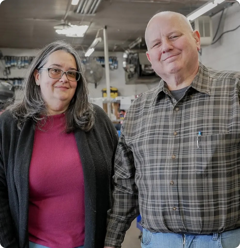 About us, auto repair in Springfield, OR, at Absolute Air & Auto Repair. A woman in a red shirt and black cardigan stands next to a man in a plaid shirt and jeans, both smiling inside a workshop or garage with tools and equipment in the background.