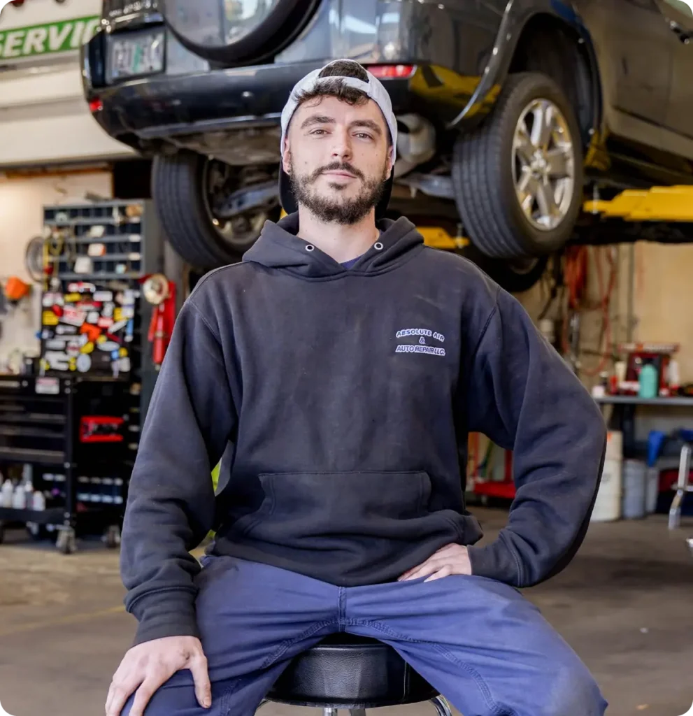 Career, auto repair in Springfield, OR, at Absolute Air & Auto Repair. A mechanic in a black hoodie and cap sits on a stool in an auto repair shop, with one hand on his knee and a car lifted on a hydraulic lift in the background. Tools and equipment are visible around the workshop.