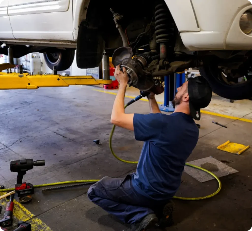 Vehicles we service, auto repair in Springfield, OR, at Absolute Air & Auto Repair. A mechanic wearing a cap kneels on the ground while repairing the exposed brake system of a white car lifted on a hydraulic lift in an auto repair shop. Tools and equipment are visible nearby.