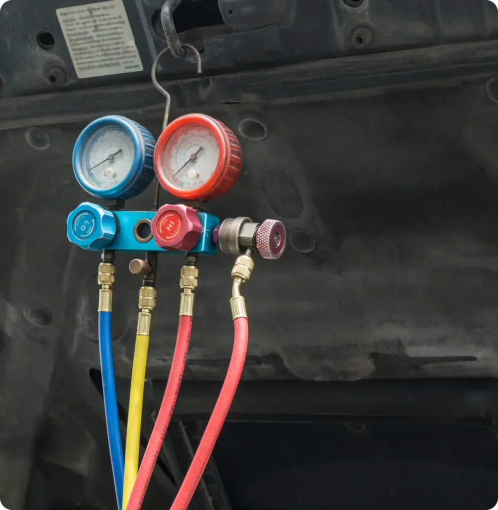 A close-up of a manifold gauge set with blue, red, and yellow hoses used for diagnosing and servicing air conditioning systems, hanging under an open car hood.