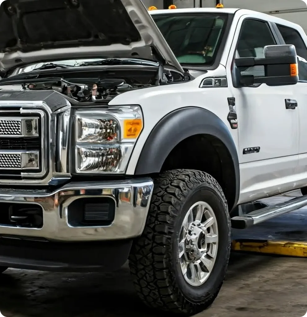 A white pickup truck with its hood open is parked indoors on a vehicle lift, indicating it is undergoing maintenance or repairs.
