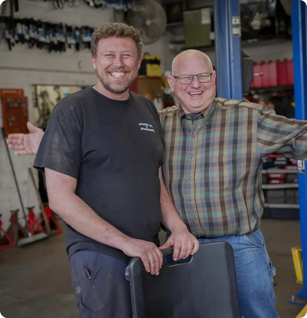 Two men smiling and posing together in an auto repair shop; one is wearing a black shirt with visible grease stains, and the other wears glasses and a plaid shirt. Car repair tools and equipment are visible in the background.