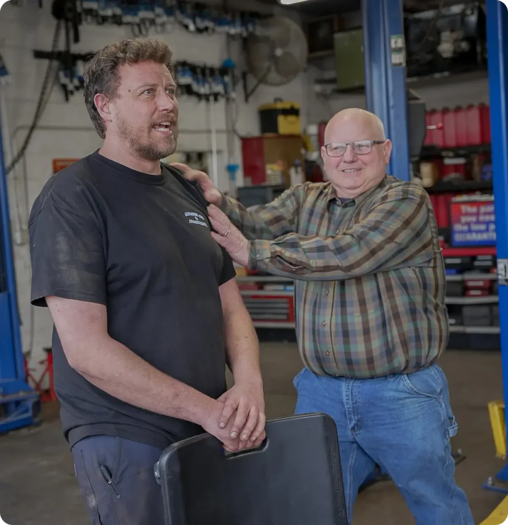 Two men in a mechanic shop; one stands with dirty hands and a black shirt, looking ahead, while the other, wearing glasses and a plaid shirt, smiles and gently places his hands on the first mans shoulders.