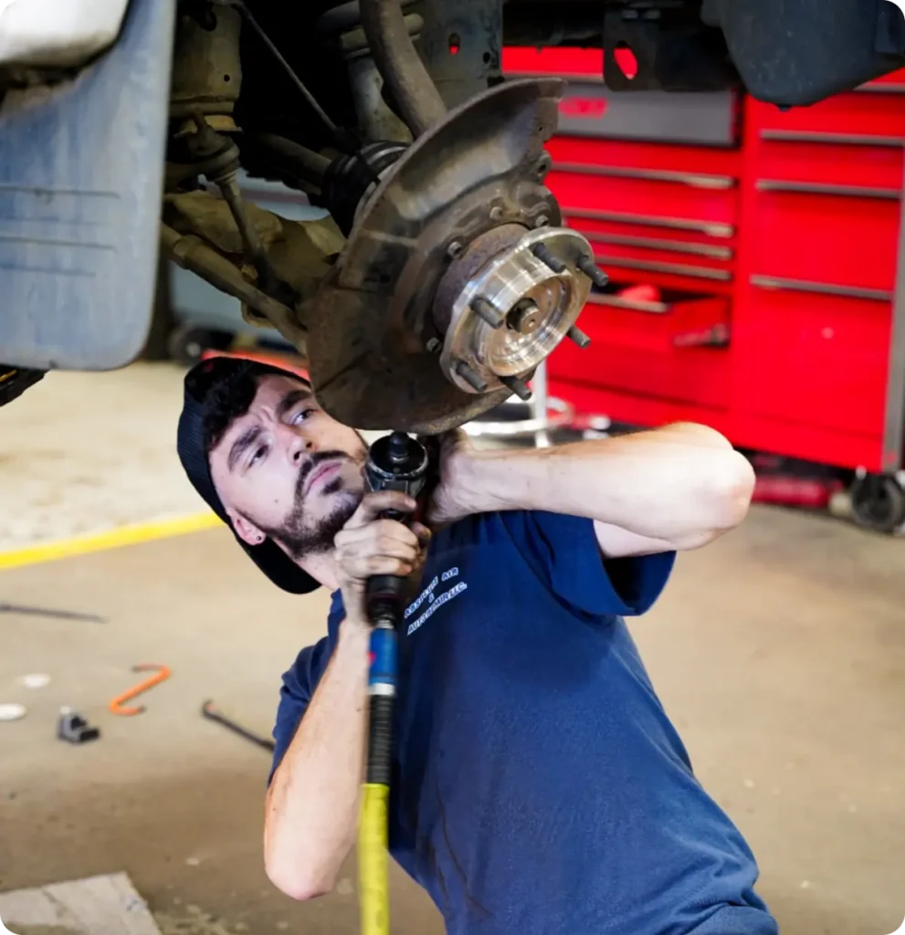 A mechanic in a blue shirt and backwards cap uses a power tool to work on the underside of a vehicle in a garage, with a red tool chest in the background.