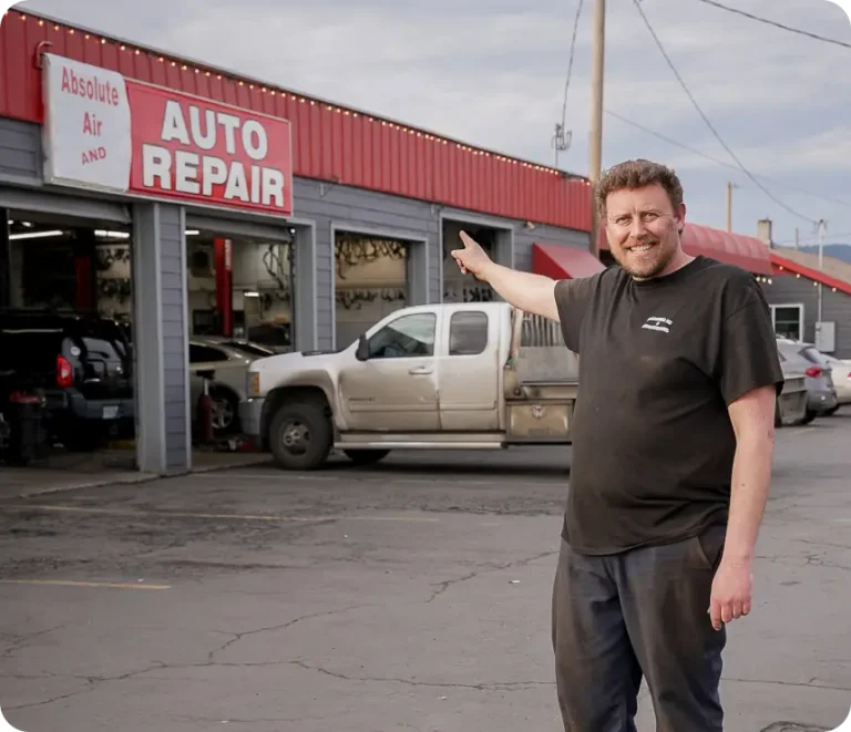 A man in a black shirt stands in front of an auto repair shop, smiling and pointing toward a large AUTO REPAIR sign on the building, with trucks parked nearby.