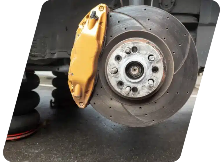 Close-up of a cars exposed brake disc and caliper, with the wheel removed. The brake caliper is painted gold, and the ventilated brake disc shows drilled holes and wear. The image is taken at an angle.
