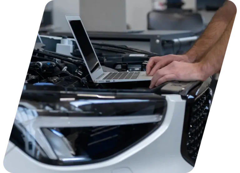 A person uses a laptop on the hood of a white car with the hood open, likely diagnosing or repairing the vehicle. Only their hands and forearms are visible.