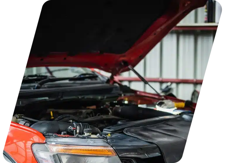 A red car with its hood open inside a garage or workshop, exposing the engine and parts, with a black cover protecting the front fender.