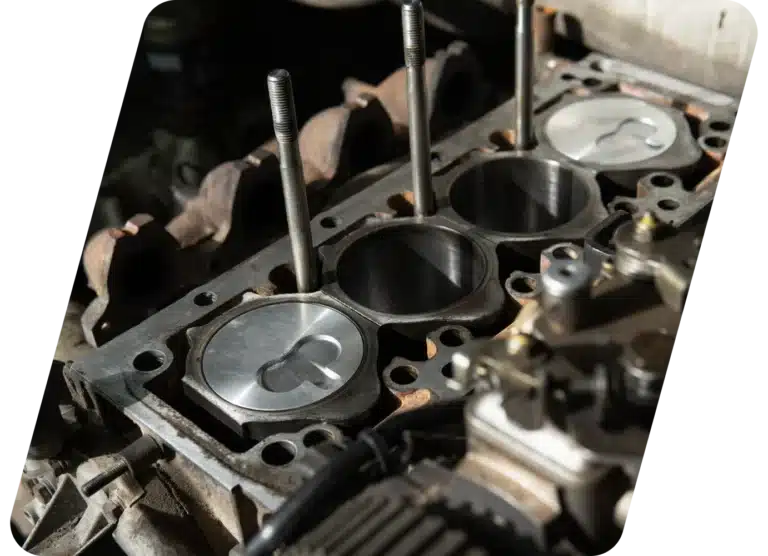 Close-up of an exposed engine block with visible pistons and cylinder walls, partially disassembled, showing intricate metal parts and components in a workshop setting.