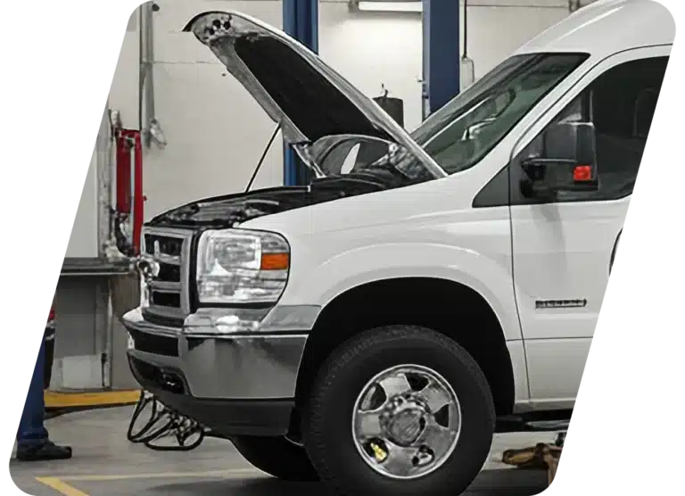 A white van with its hood open is parked inside an auto repair shop, with cables connected to the front.