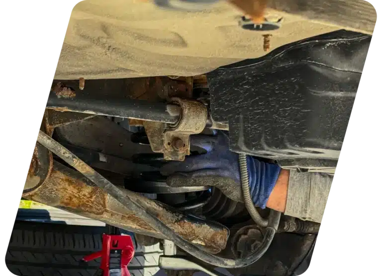 A person wearing a blue glove works underneath a vehicle, handling suspension components near a black fuel tank. A jack stand and a tire are visible in the background.