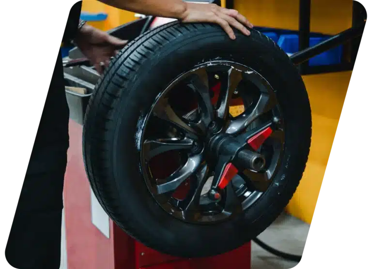 A person is holding a car tire mounted on a red machine, likely balancing or installing the tire in an auto repair shop with yellow and blue background elements.