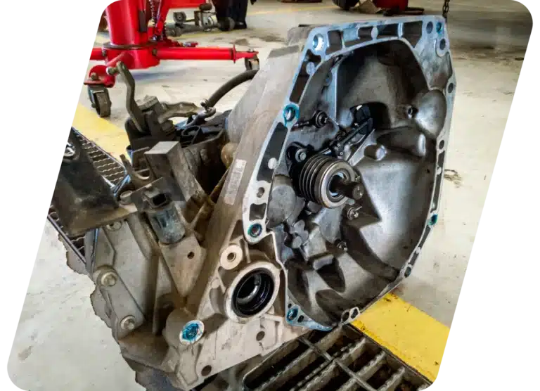 A close-up of a car transmission placed on a workshop floor, showing gears and components inside the metal housing, with workshop equipment visible in the background.