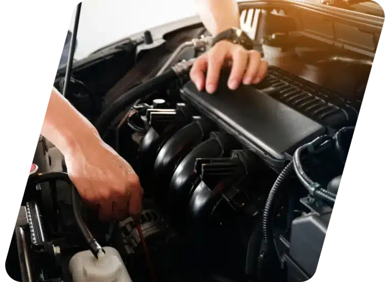 A person’s hands working on a car engine, with one hand holding a tool and the other resting on a black engine cover, showing an automotive maintenance or repair scene.